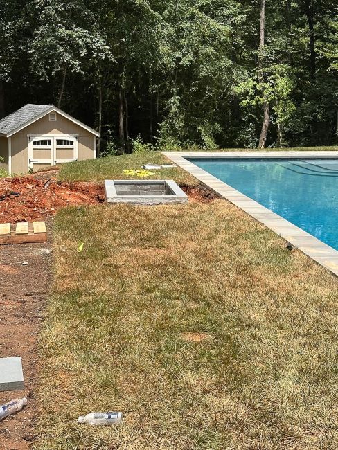 A newly constructed rectangular concrete underground TV lift vault next to a backyard swimming pool under construction, with a storage shed in the background.