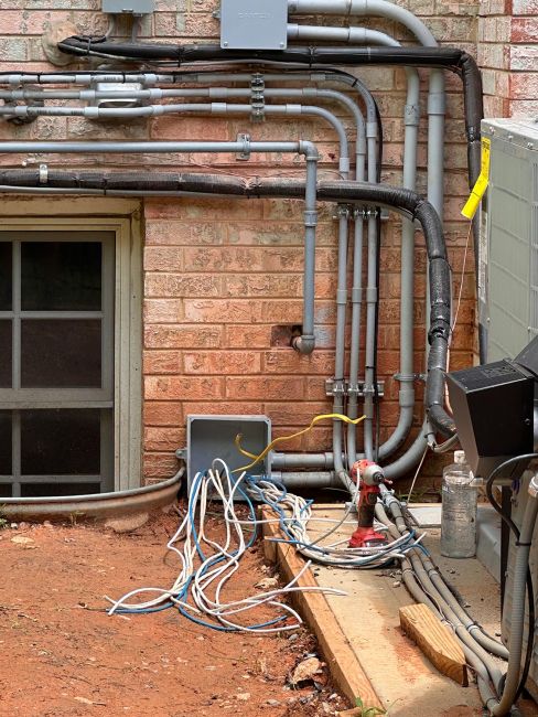 Complex outdoor electrical wiring, conduits, and an open junction box with exposed cables on a brick wall next to an HVAC unit.