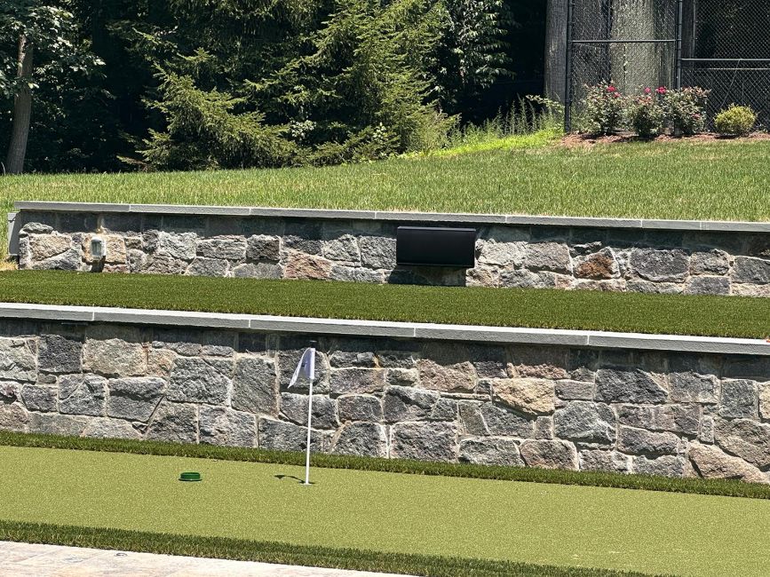 A black weather-resistant outdoor speaker mounted flat against a stone retaining wall directly above a backyard putting green.