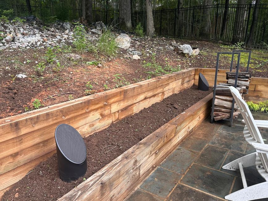 Two sleek, dark grey cylindrical outdoor landscape speakers installed in a freshly mulched garden bed behind a wooden retaining wall.