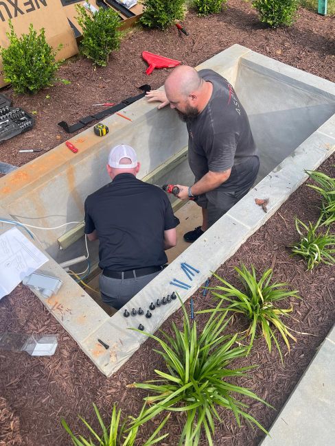 Two technicians working inside a concrete underground bunker to install a motorized outdoor TV lift system, surrounded by tools and mulch.