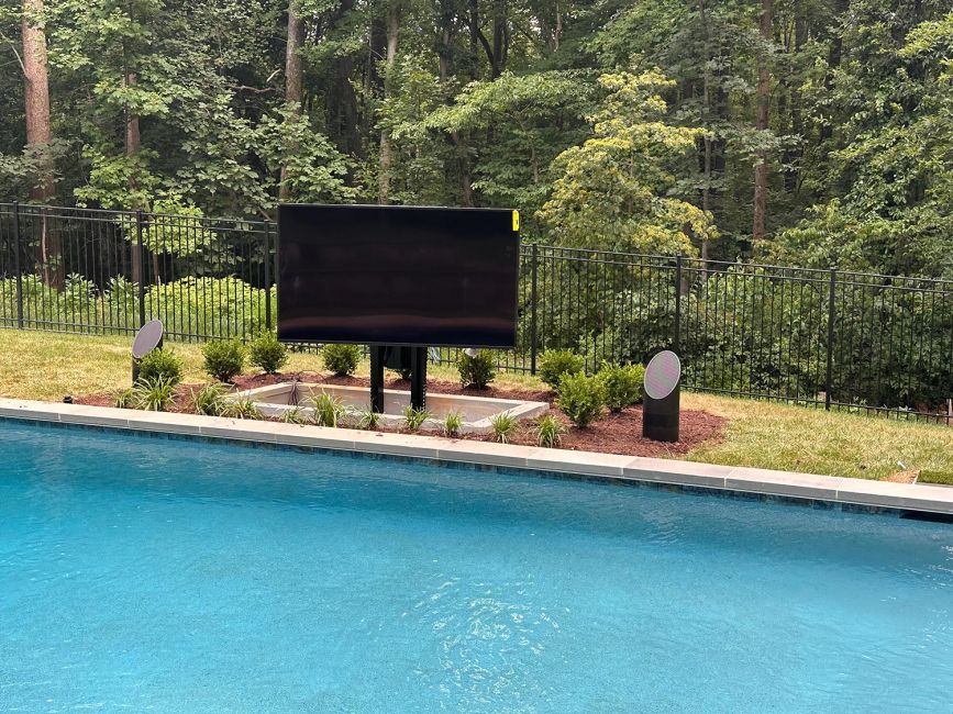 A large flat-screen outdoor television fully raised from its hidden underground vault, positioned next to a luxury backyard swimming pool.