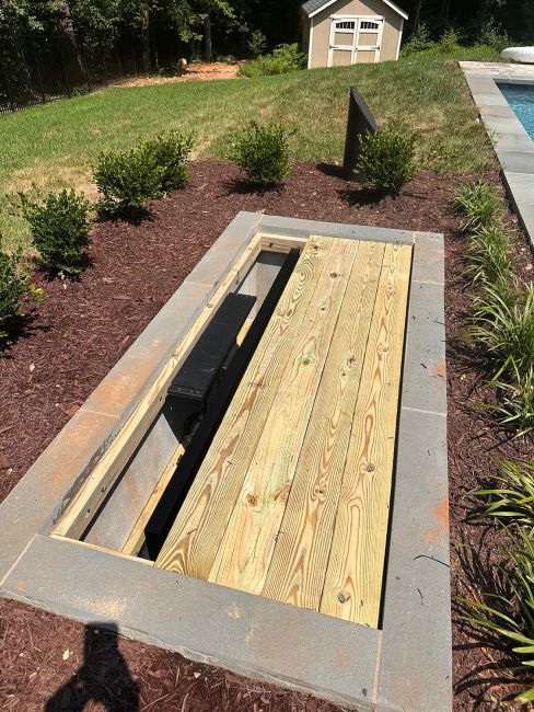 The hidden outdoor TV lift concrete vault partially covered with a custom wooden plank lid, flush with the surrounding garden mulch and landscaping.